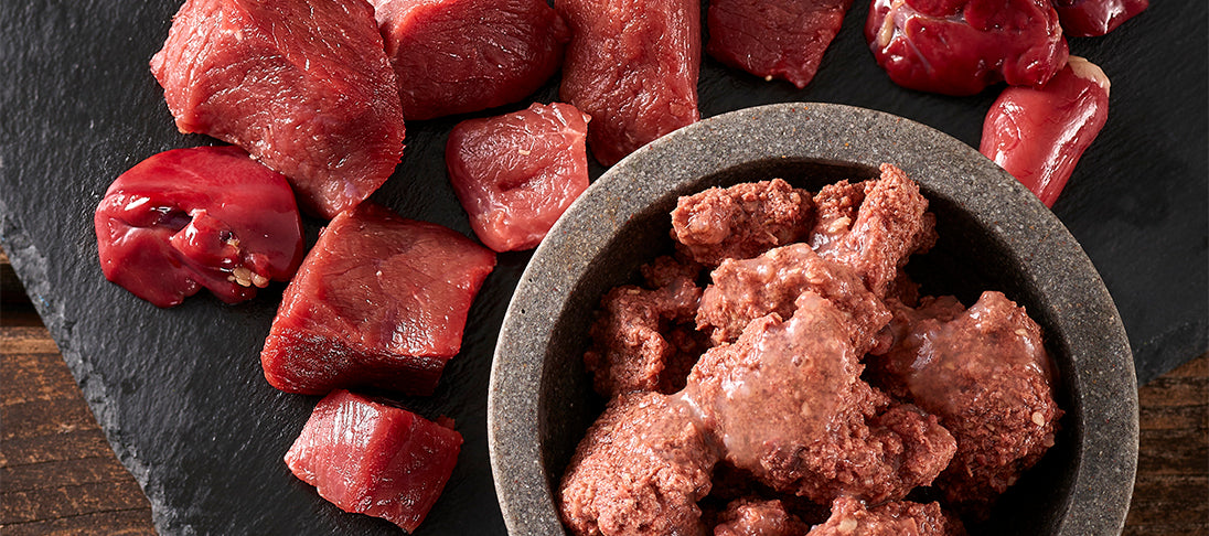 Auf einer Platte liegen rohe Fleischstücke und Innereien sowie eine Schale mit der Reinfleischlinie Steakhouse von Fleischeslust für Nass-BARF.